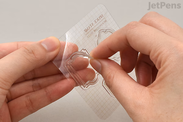 A person peeling a clear acrylic stamp from a transparent backing sheet, holding it between their fingers. The stamp is shaped like a flower.
