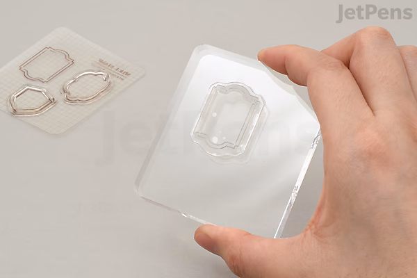 A hand holding a clear square acrylic block with a rectangular indentation in the center, used for mounting clear stamps. Two small clear stamps with geometric designs are placed on a flat surface nearby.