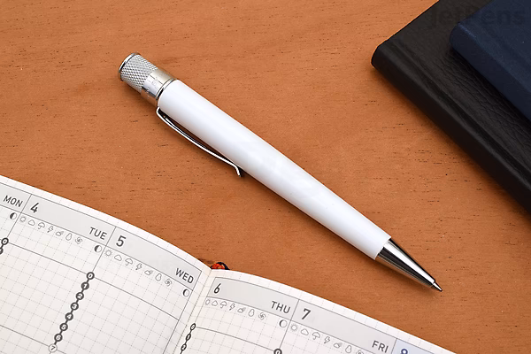 A Retro 51 Tornado Classic Lacquers rollerball pen with a sleek black body and silver accents, placed on a brown surface next to an open planner and a closed notebook. The pen has a 0.7 mm tip.