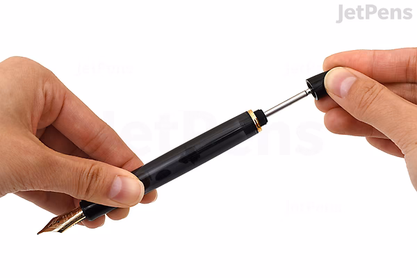 A person holding a Pilot Custom 823 fountain pen in amber brown, demonstrating the removal or insertion of the pen's ink converter. The pen has a transparent barrel and a gold-colored nib.