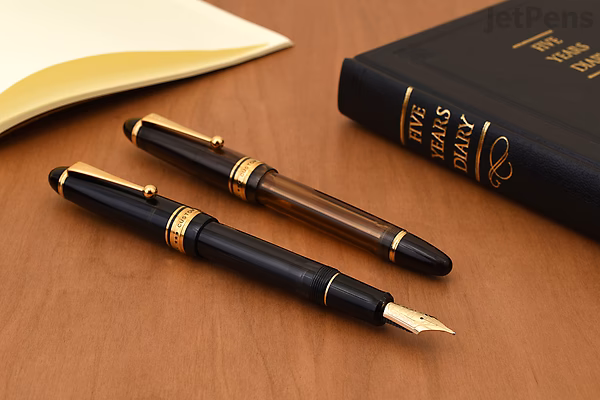 Two elegant black and gold Pilot Custom 823 fountain pens, one with the cap on and one uncapped showing the gold nib, displayed on a wooden surface next to a closed black book and a cream-colored item.