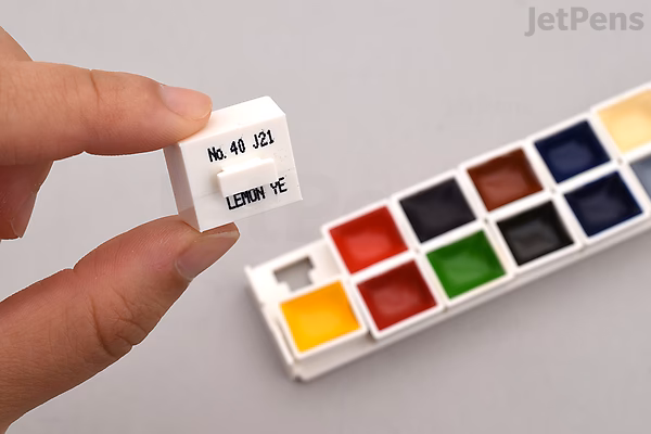 A hand holding a small white label with Japanese text and numbers next to a 14-color watercolor palette featuring vibrant shades including yellow, red, green, blue, and black.