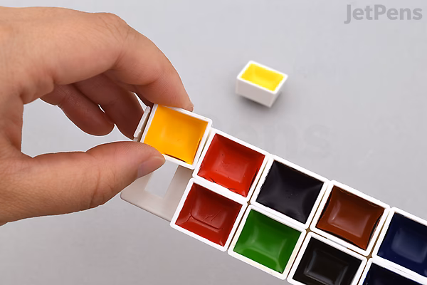 Hand holding a yellow watercolor pan above a white plastic palette containing multiple watercolor pans in various colors.