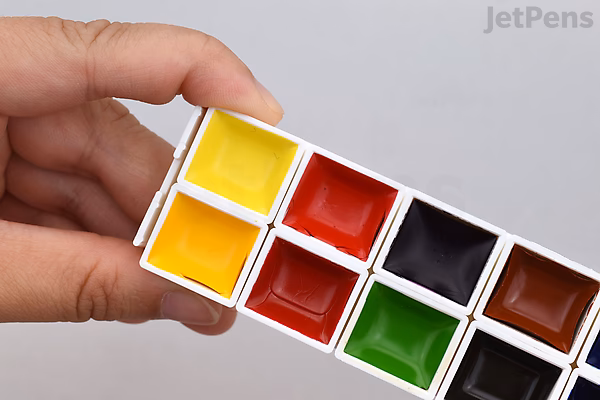 A hand holding a small watercolor palette containing 14 vibrant paint colors arranged in two rows, including yellow, red, black, green, and brown shades. The palette is labeled as Kuretake Gansai Tambi Mini Watercolor Palette.