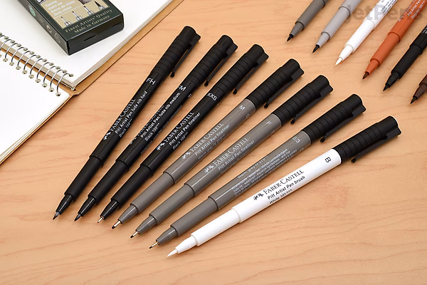 A set of eight Faber-Castell PITT Artist Pens arranged in a row on a wooden surface, featuring various black pen tips and barrel designs, alongside some sketching tools and a notebook in the background.