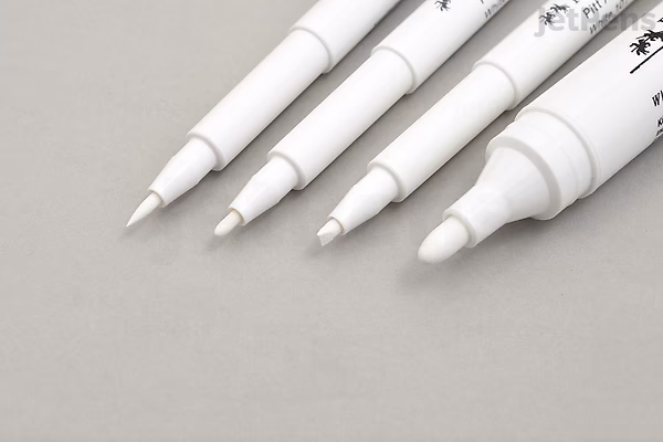 Four white Faber-Castell PITT Artist Pens arranged side by side on a gray surface, showing different tip sizes and shapes from fine brush to broad marker.