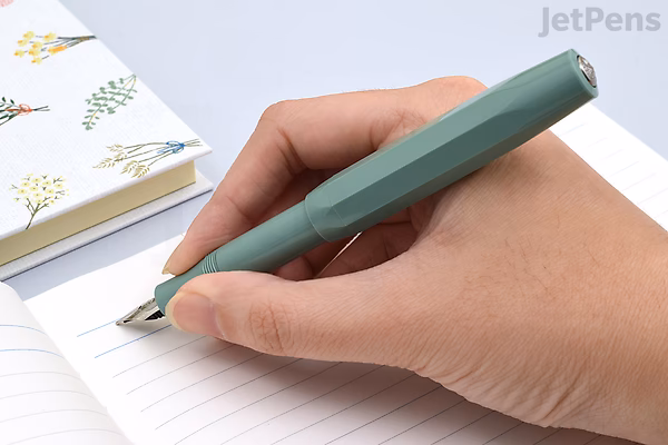 A hand holding a Kaweco Collection Sport Fountain Pen in Smooth Sage color with a medium nib, writing on a lined notebook. The pen has a compact, hexagonal design and a matte finish. A floral-patterned notebook is partially visible in the background.