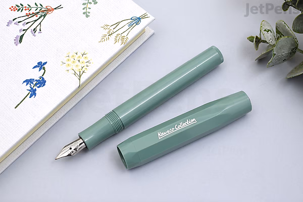 A Kaweco Collection Sport Fountain Pen in smooth sage green with a medium nib, shown uncapped next to its cap on a light gray surface. A floral-patterned notebook and a small green plant are partially visible nearby.