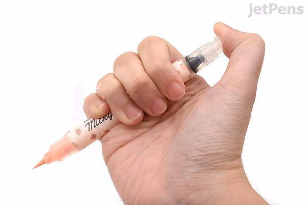 A hand holding a Pentel Milky Brush Pen with a light peach-colored tip, shown against a white background. The pen has a clear barrel with visible ink inside and black text printed on it.