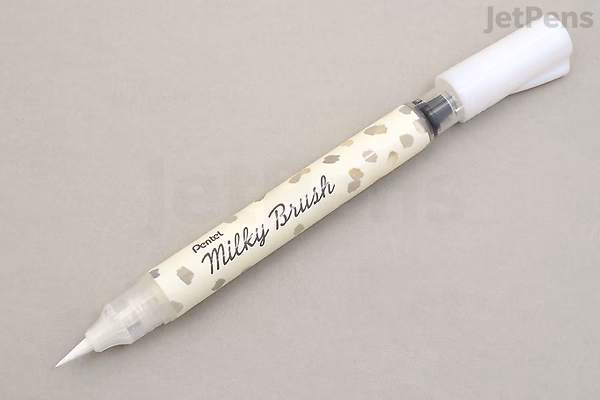 A Pentel Milky Brush Pen in white, featuring a clear barrel with a white brush tip and a white cap, lying on a light gray surface. The pen has a label with the text "Milky Brush" and small decorative dots.