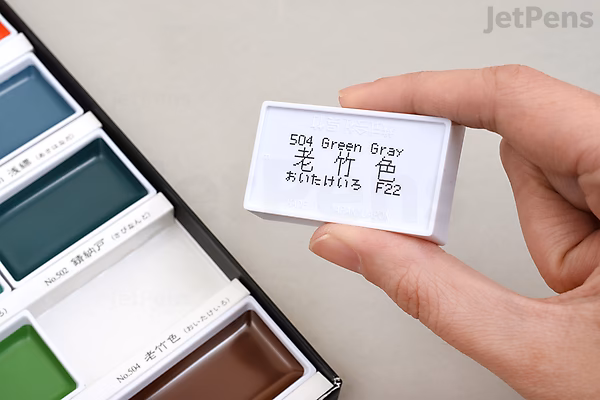 Hand holding a small white box labeled "504 Green Gray" with Japanese characters and numbers, next to an open watercolor palette with various colors.