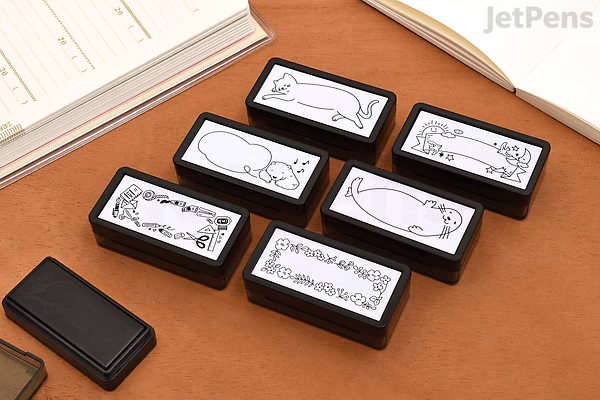 A set of seven rectangular black pre-inked stamps with white labels showing various bird illustrations and decorative designs, arranged on a brown surface next to a notebook.