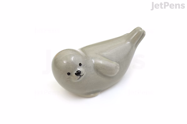 A small, smooth ceramic pen rest shaped like a gray seal lying on its belly with a simple, smiling face and flippers.