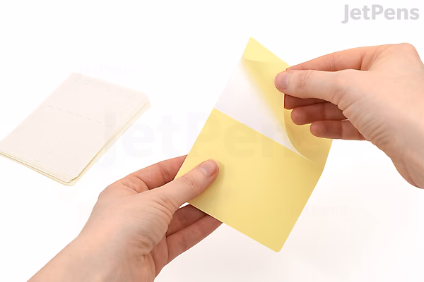 A person peeling a yellow removable calendar sticker from its backing sheet. Another sheet of stickers is visible in the background on a white surface.