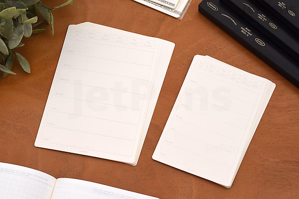 Two sheets of small, undated weekly calendar stickers with horizontal lines for writing, placed on a wooden surface next to a plant and a black pen case.