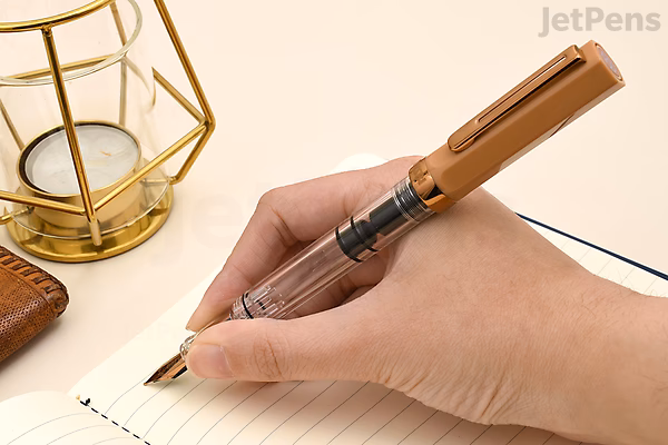 A hand holding a TWSBI ECO Caffè Bronze fountain pen with a 1.1 mm stub nib, writing on a lined notebook. The pen has a bronze-colored body and a transparent section showing the ink inside. A geometric gold candle holder and a brown leather item are visib