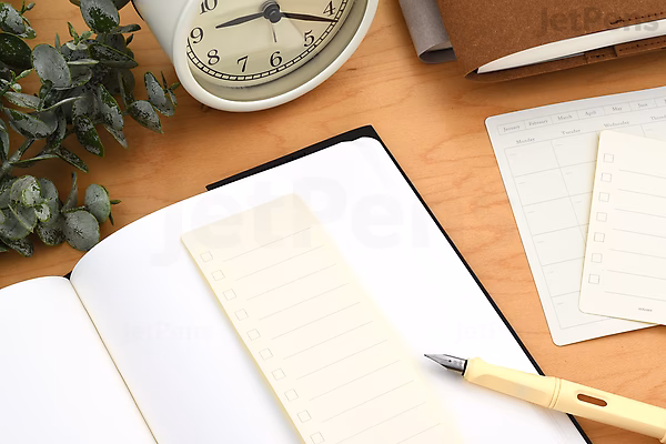 A flat lay of a workspace featuring an open notebook with lined removable calendar stickers placed on its pages. Nearby are additional calendar stickers, a beige pen, a round clock showing 10:10, and a small green plant on a wooden surface.