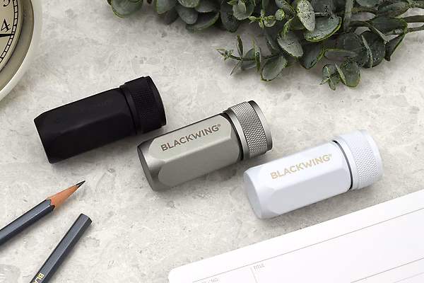 Three Blackwing One-Step Long Point Pencil Sharpeners in black, grey, and white are arranged on a light surface next to two sharpened pencils and a plant.