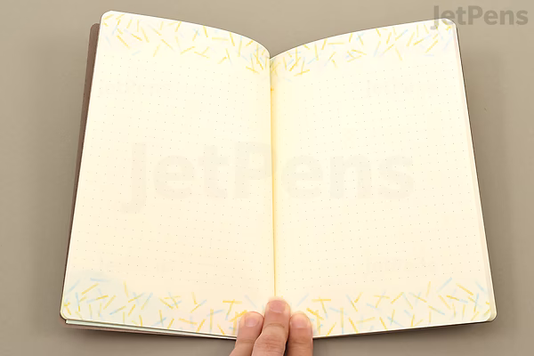 Open Midori Yuru Log Notebook in B6 size with cream-colored dotted pages and a subtle watercolor floral border at the top and bottom edges. A hand is holding the notebook at the bottom center.
