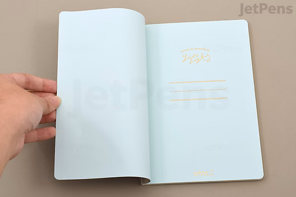 A hand is holding open a gray Midori Yuru Log Notebook (B6 size) showing a free monthly diary page with simple yellow printed text and lines on a light blue page. The notebook has thick MD paper.
