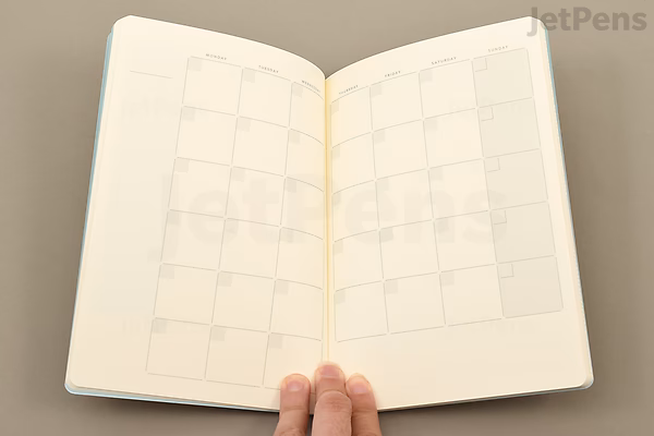 Open notebook showing a two-page monthly calendar layout with blank square date boxes, held by a hand at the bottom center. The pages are cream-colored with light gray lines.