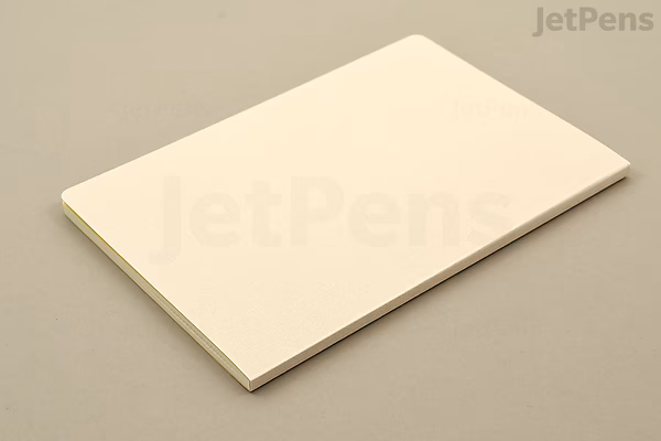 A closed beige notebook with a simple, clean cover, shown lying flat on a light surface. The notebook is a Midori Yuru Log Notebook in B6 size with MD Thick paper and 2 mm grid pages.