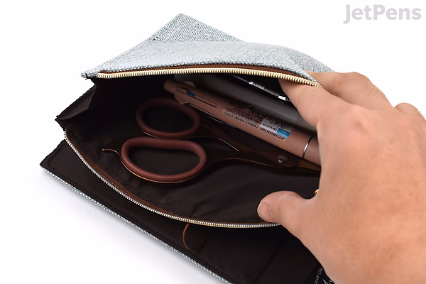 Beige cloth zipper pouch with a B6 notebook cover design, partially open to reveal interior pockets holding scissors and pens, held by a hand.