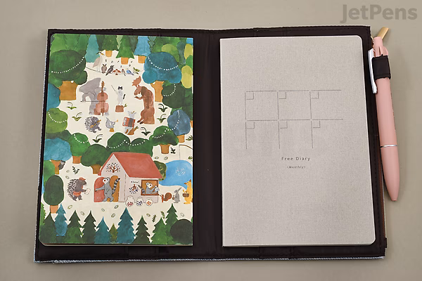 Open beige cloth notebook cover designed for B6 size, featuring a colorful illustrated zipper pouch on the left with a whimsical village scene, and a plain notebook on the right with a simple grid layout. A pink pen is clipped to the right side.