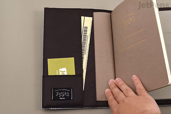 Open beige cloth notebook cover with multiple interior pockets, including a black pocket holding papers and a green card, designed to fit a B6 notebook. A hand is holding the cover open.