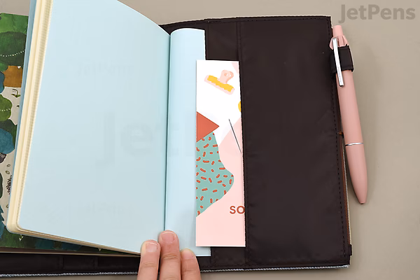 Beige cloth zipper pouch notebook cover in B6 size, holding an open notebook with light blue pages, a colorful bookmark, and a pink pen clipped to the side.