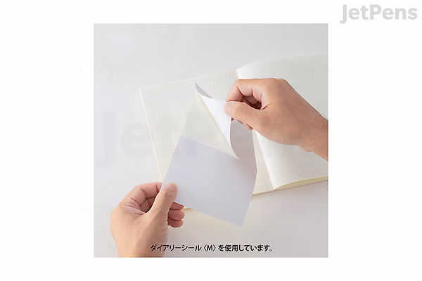 A person peeling a transparent sticker from a white backing sheet, holding it above an open Midori MD Diary 2025 planner. The scene shows a close-up of hands and the planner on a white surface.