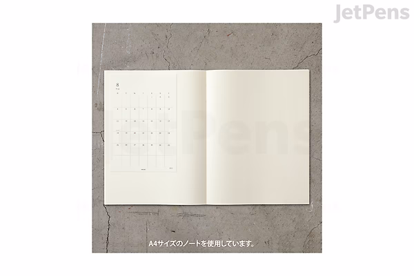 An open 2025 Midori MD diary showing a monthly calendar on the left page and a blank page on the right, placed on a textured gray surface.