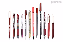 A lineup of twelve red and black pens and pencils arranged horizontally on a white background, showcasing various styles and brands from the JetPens Red Black Pen Sampler.