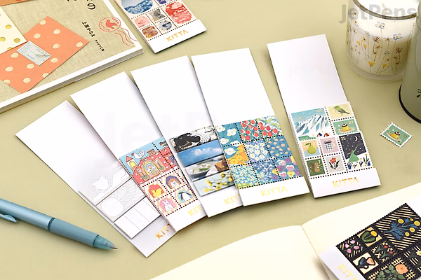 Five packs of King Jim Kitta Washi Tape, each with colorful and patterned tape samples displayed on white cards, laid out on a light surface next to a blue pen, an open notebook, and other stationery items.