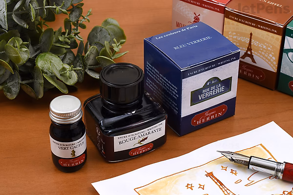 Bottles of Herbin Ambre de Birmanie ink, including a 10 ml bottle and a larger bottle with its box, displayed on a wooden surface with a fountain pen and paper nearby.