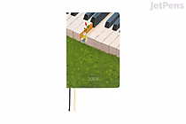 Hobonichi Techo A6 planner cover illustration: person sitting on oversized piano keys above a green grassy field with "2026" printed on the grass; two thin ribbon bookmarks.